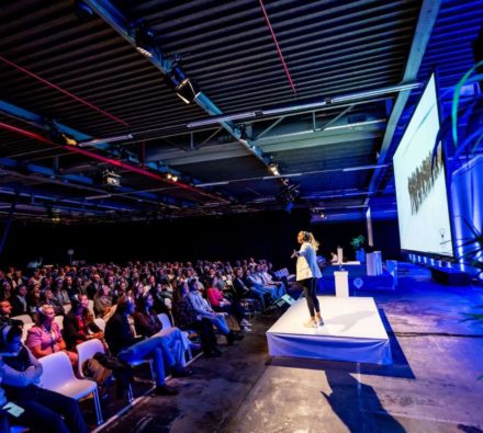 Spreker op een podium voor een volle zaal tijdens een congres