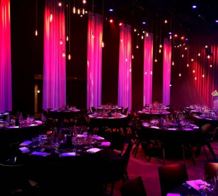 Ronde dinertafels in een zaal met roze en paarse verlichting en hoge gordijnen bij Hart van Holland